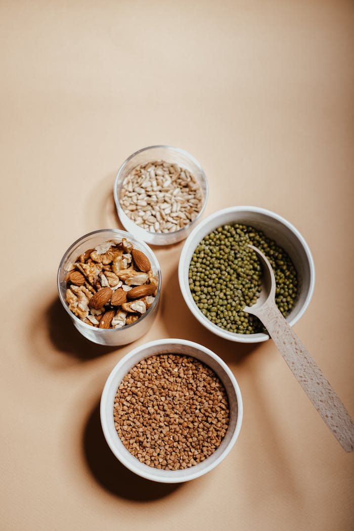 An overhead view of assorted nuts and grains in bowls with a spoon on a neutral background.